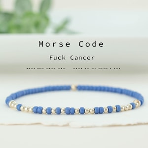May include: A blue beaded bracelet with gold-colored beads. The text "Morse Code Fuck Cancer" is above the bracelet. The bracelet is on a white surface with a blurred green background.