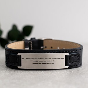 May include: A black leather bracelet with a silver plate engraved with braille dots.