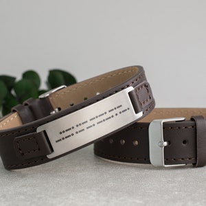 Pode incluir: Duas pulseiras de couro marrom com fivelas prateadas. Uma pulseira tem uma placa prateada com uma mensagem em braille.