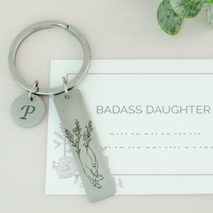 May include: Silver keychain with a small round charm engraved with the letter "P" and a rectangular charm with an engraved design of a hand holding flowers. The rectangular charm has the text "BADASS DAUGHTER" engraved on it.