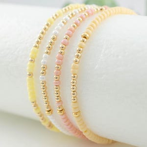 May include: Four beaded bracelets in soft yellow, pearl white, tea rose pink, and pear colors. Each bracelet features small, round beads and gold-colored accents. The bracelets are stacked together, showcasing their delicate design.