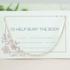 May include: A silver beaded bracelet with the text "I'd help bury the body" on a white card with pink floral accents.