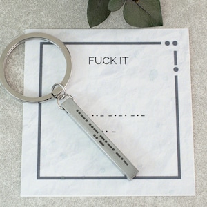 Op de afbeelding: Een zilverkleurige sleutelhanger met een rechthoekige hanger gegraveerd met morsecode. De sleutelhanger is bevestigd aan een grote metalen ring. De achtergrond toont een witte kaart met de woorden "FUCK IT" en een decoratieve rand.