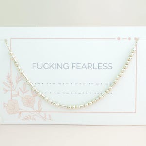 May include: A silver beaded necklace displayed on a white card with the text "FUCKING FEARLESS". The card features a pink floral design and border. The necklace is curved and delicate. The background is a soft, neutral colour.