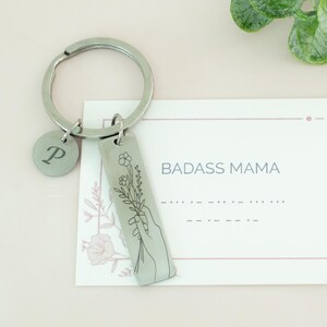 May include: Silver keychain with a floral design and the words "BADASS MAMA" on a white card. The keychain has a letter "P" charm.