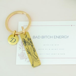 Op de afbeelding: Gouden sleutelhanger met een bedel waarop "BAD BITCH ENERGY" en een bloemenmotief staat. De sleutelhanger heeft ook een kleine bedel met de letter "D" erop.