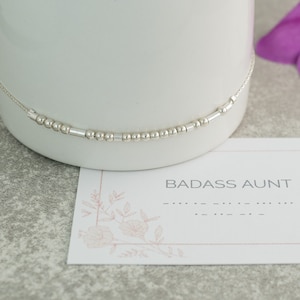 May include: A silver beaded bracelet with the words "BADASS AUNT" and braille lettering on a white card with a pink floral design.