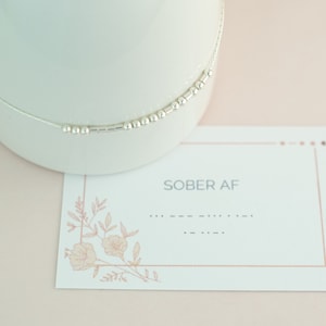May include: A silver beaded necklace rests on a white ceramic surface, next to a card that reads "SOBER AF" in gray text. The card has a floral design in the corner and a dotted line. The background is a soft pink.