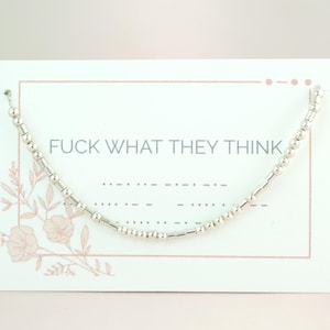 Op de afbeelding: Een zilveren kralenketting in een gebogen vorm, gepresenteerd op een witte kaart. De kaart heeft de tekst "FUCK WHAT THEY THINK" in het zwart, met een bloemenontwerp. Het is een sieraad.