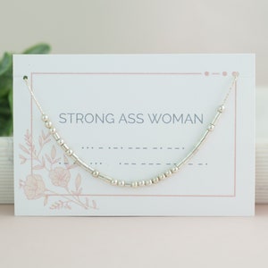 May include: A silver beaded necklace with the text "STRONG ASS WOMAN" on a white card with pink floral accents.