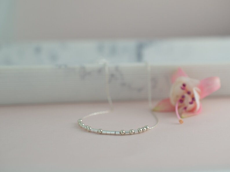 May include: A delicate silver necklace with a series of small, round beads and cylindrical bars. The necklace is displayed on a soft pink surface, with a blurred pink flower in the background. The necklace is a minimalist piece of jewelry.