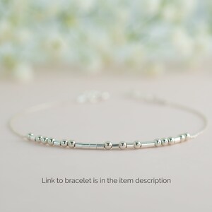 May include: A delicate silver bracelet with small round beads and a thin chain. The bracelet is on a light pink background.