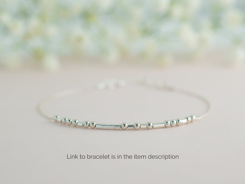 May include: A delicate silver bracelet with small round beads and a thin chain. The bracelet is on a light pink background.