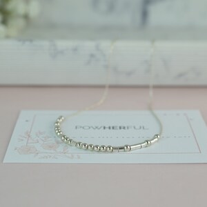 May include: A silver necklace with small round beads and silver bars spelling out the word "powerful" on a white card with a floral design.