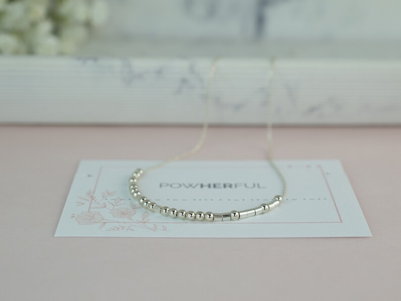 May include: A silver necklace with small round beads and silver bars spelling out the word "powerful" on a white card with a floral design.