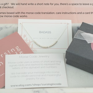 May include: A close-up of a small, white card with the word "BADASS" printed in black. A silver necklace with small beads is displayed on the card. The necklace is spelling out the word "BADASS" in Morse code. The card is sitting on a pink surface with the text "cursingincode" printed on it.