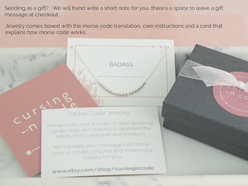 May include: A close-up of a small, white card with the word "BADASS" printed in black. A silver necklace with small beads is displayed on the card. The necklace is spelling out the word "BADASS" in Morse code. The card is sitting on a pink surface with the text "cursingincode" printed on it.