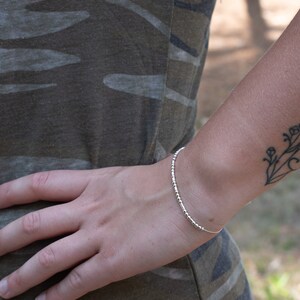 May include: A silver beaded bracelet on a person's wrist. The bracelet is thin and delicate, with small, evenly spaced beads. The person is wearing a camouflage shirt.