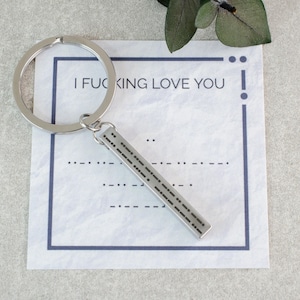Può includere: Un portachiavi argentato con un ciondolo argentato che dice "I F**KING LOVE YOU" in braille. Il portachiavi è su una carta bianca con un bordo blu.