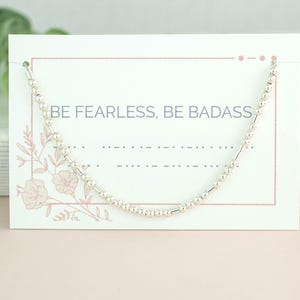 Pode incluir: Um bracelete de contas prateadas com o texto "BE FEARLESS, BE BADASS" em um cartão branco com desenhos florais rosa.