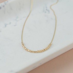 May include: A delicate gold necklace featuring a series of small, round beads and cylindrical bars strung along a fine chain. The necklace is displayed on a white marble surface, showcasing its minimalist design.