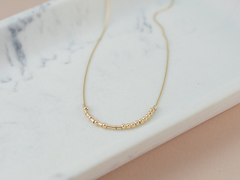 May include: A delicate gold necklace featuring a series of small, round beads and cylindrical bars strung along a fine chain. The necklace is displayed on a white marble surface, showcasing its minimalist design.