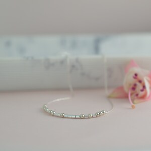 May include: A delicate silver necklace with small round beads and a simple design. The necklace is displayed on a pink surface with a blurred background.