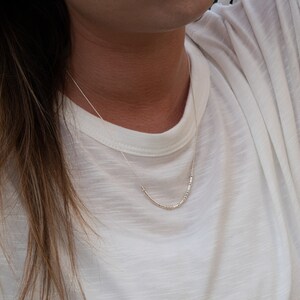 May include: A delicate silver necklace with a curved bar pendant. The necklace is worn against a white t-shirt. The pendant has a series of small, evenly spaced beads. The necklace chain is thin and subtle.