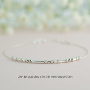 May include: A delicate silver bracelet with small round beads and a thin chain. The bracelet is on a light pink background.