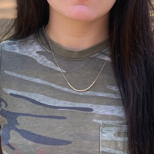 May include: A delicate gold necklace featuring a series of small, round beads strung along a fine chain. The necklace is worn against a camouflage-patterned shirt, creating a contrast between the jewelry and the casual attire.