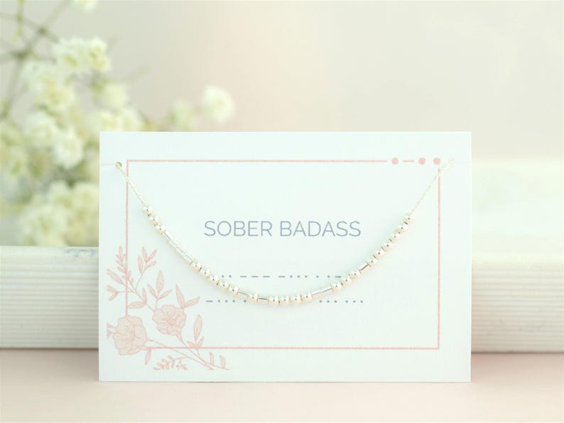 May include: A white card with pink floral accents and the text "SOBER BADASS" printed on it. A silver beaded necklace is displayed on the card.