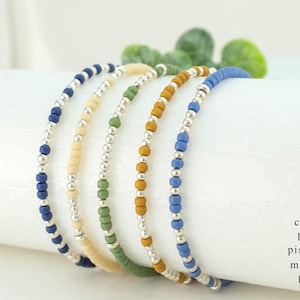 May include: Five beaded bracelets in shades of blue, yellow, green, and mustard. Each bracelet features small, colorful beads interspersed with silver-colored beads. The bracelets are displayed on a white surface, with the color names listed in the image.