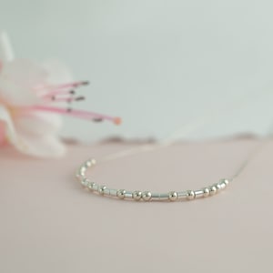 May include: A delicate silver necklace with small round and rectangular beads. The necklace is displayed on a pink surface.