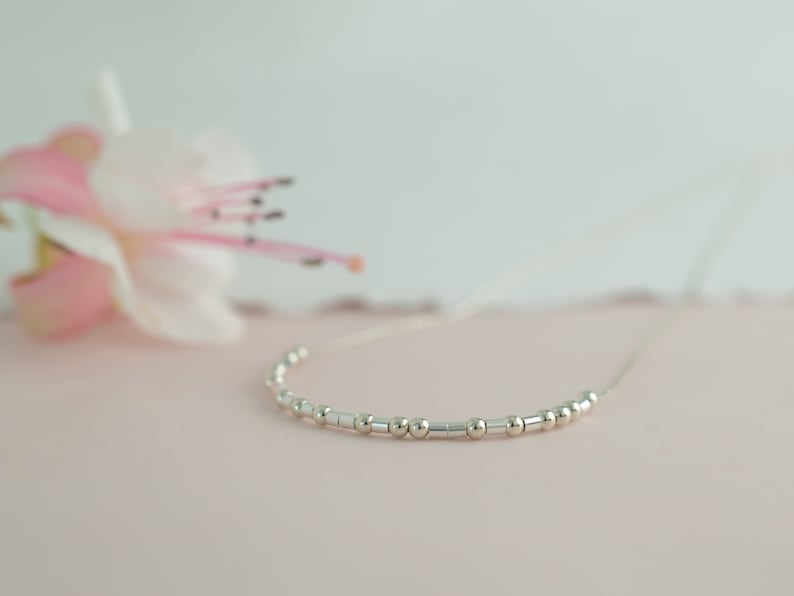 May include: A delicate silver necklace with small round and rectangular beads. The necklace is displayed on a pink surface.