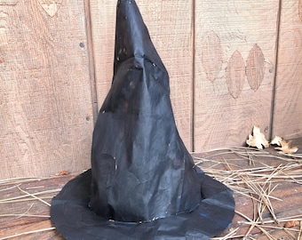 May include: A black witch hat with a wide brim and a tall, pointed crown. The hat is made of a dark, textured material and sits on a wooden surface with pine needles and leaves. A Halloween-themed accessory.