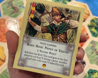 May include: A game card featuring an illustration of Robin Hood blowing a horn. The card has text that reads "Robin Hood, Prince of Thieves" and "1 Victory Point!" The card is held in a person's hand.