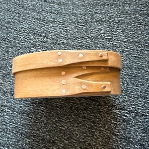 May include: A wooden oval box with a hinged lid. The box is made of light brown wood and has copper-colored metal accents around the edges.