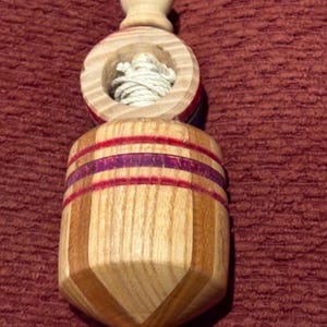 May include: A wooden top toy with a light brown knob and a round section with a white string inside. The main body is a mix of light and dark wood with purple and red stripes. The toy is designed to be spun.