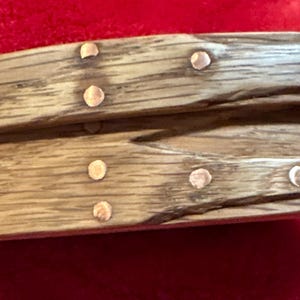 May include: A handcrafted, oval-shaped wooden box with a lid. The box is made of light-colored wood with visible grain and is secured with small, copper-colored rivets. The box is likely used for storage or decorative purposes.