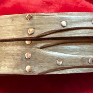 May include: A wooden oval-shaped box with a hinged lid. The box is made of two pieces of wood that are joined together with metal rivets. The box is a light brown color and has a rustic appearance.