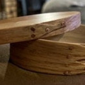 May include: A wooden oval box with a hinged lid. The box is made of light wood with a natural finish. The lid has a decorative design of woven strips and metal accents.