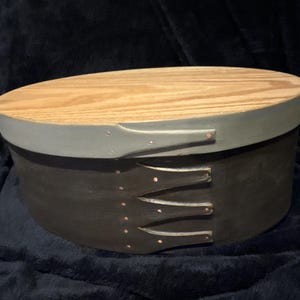 May include: Oval wooden box with a light wood lid and a dark gray body. The lid is trimmed with a light gray band. The box has three decorative handles on the side and is likely used for storage.