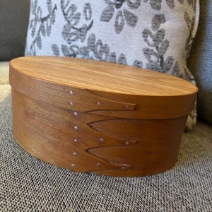 May include: Oval wooden box with a light brown finish. The box has a lid and is secured with copper rivets. The box is sitting on a gray textured surface.