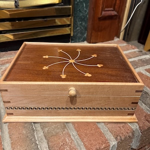 May include: A wooden box with a light-colored frame and a dark brown top. The top features a decorative floral design. The box has a small wooden knob and a decorative border. The box is sitting on a brick surface.