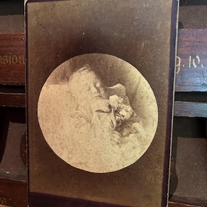 May include: Antique sepia-toned photograph of a person, possibly a child, resting peacefully. The subject is cradling flowers within a circular frame. The photograph has a dark border and is displayed at an angle, showcasing its aged appearance.
