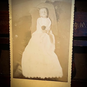 May include: A sepia-toned vintage photograph with a scalloped edge featuring a person lying down in a long, light-colored gown with a high collar. The person holds a small, dark flower in their hands. The image has a rectangular border.