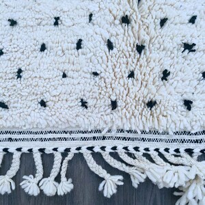 May include: White and black rug with a fringe border. The rug has a fluffy texture and a geometric pattern.