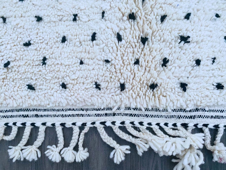 May include: White and black rug with a fringe border. The rug has a fluffy texture and a geometric pattern.