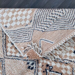 May include: Beige and cream-colored woven rug with geometric patterns. The rug features a diamond and checkered design with black accents. The rug is folded over, revealing the underside with fringe.