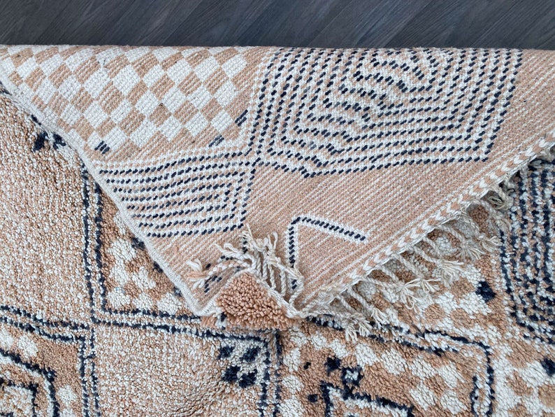May include: Beige and cream-colored woven rug with geometric patterns. The rug features a diamond and checkered design with black accents. The rug is folded over, revealing the underside with fringe.